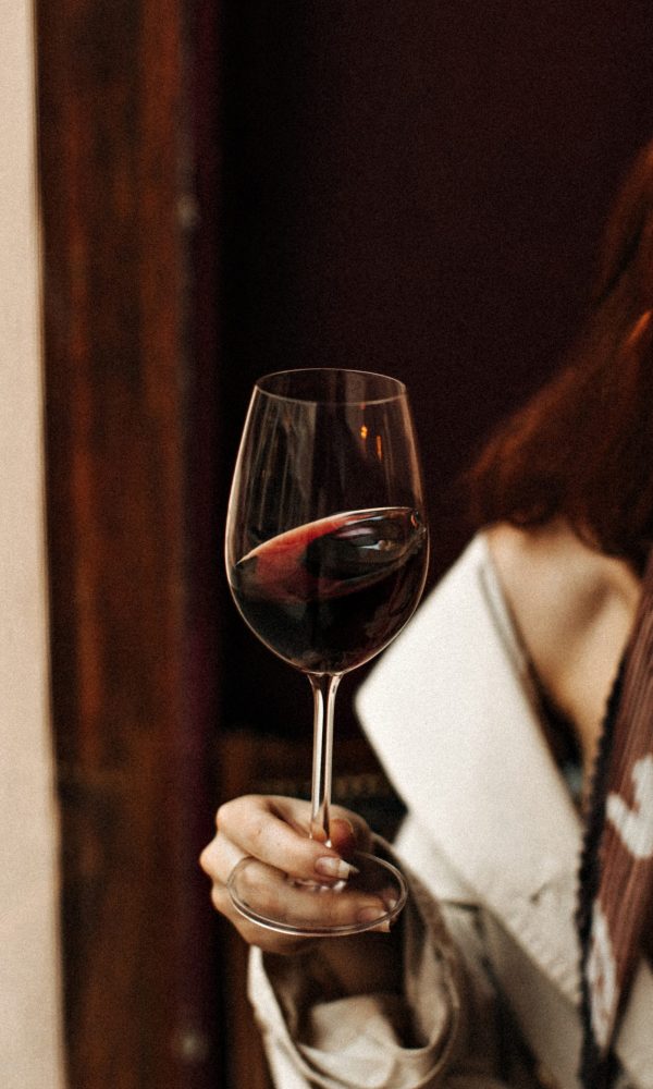 Snapshot of wine glass. Red-haired woman in white dress poses with alcohol in bar